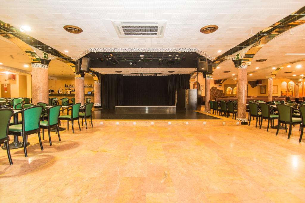 Dejotāju skatuve ar melnu aizkaru un zaļām krēsliem, kas izvietoti ap plašo zāli. Telpa ir rotāta ar kolonnām un siltu apgaismojumu, radot viesmīlīgu atmosfēru.