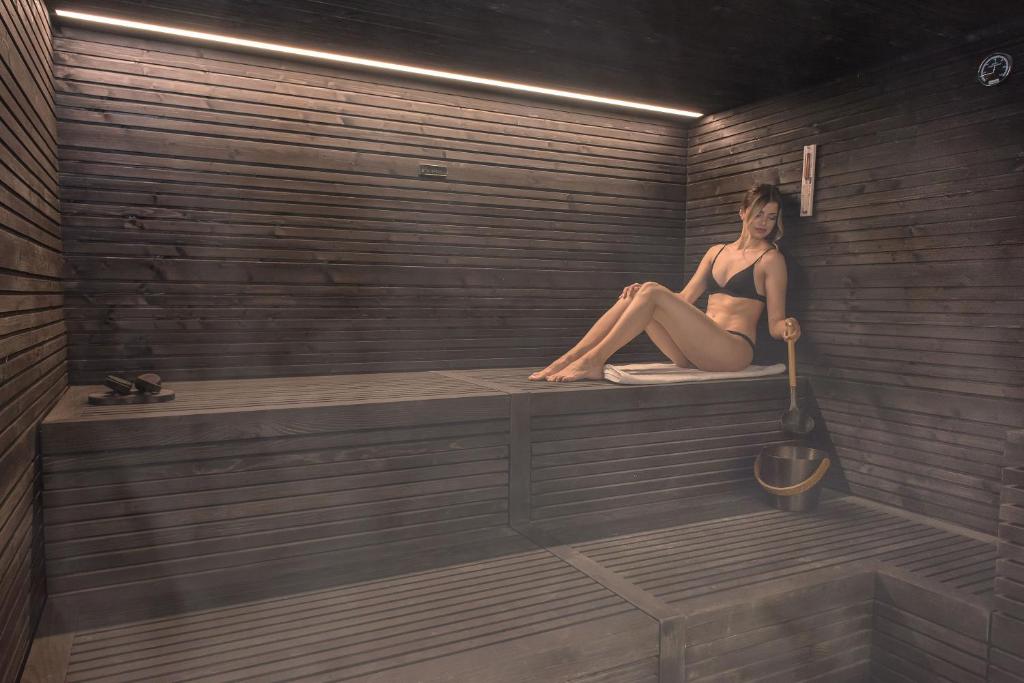 Mūsdienīga sauna ar koka apdari, kurā relaksējas sieviete. Apkārt ir tvaiks un saunas piederumi, radot mierīgu atmosfēru.