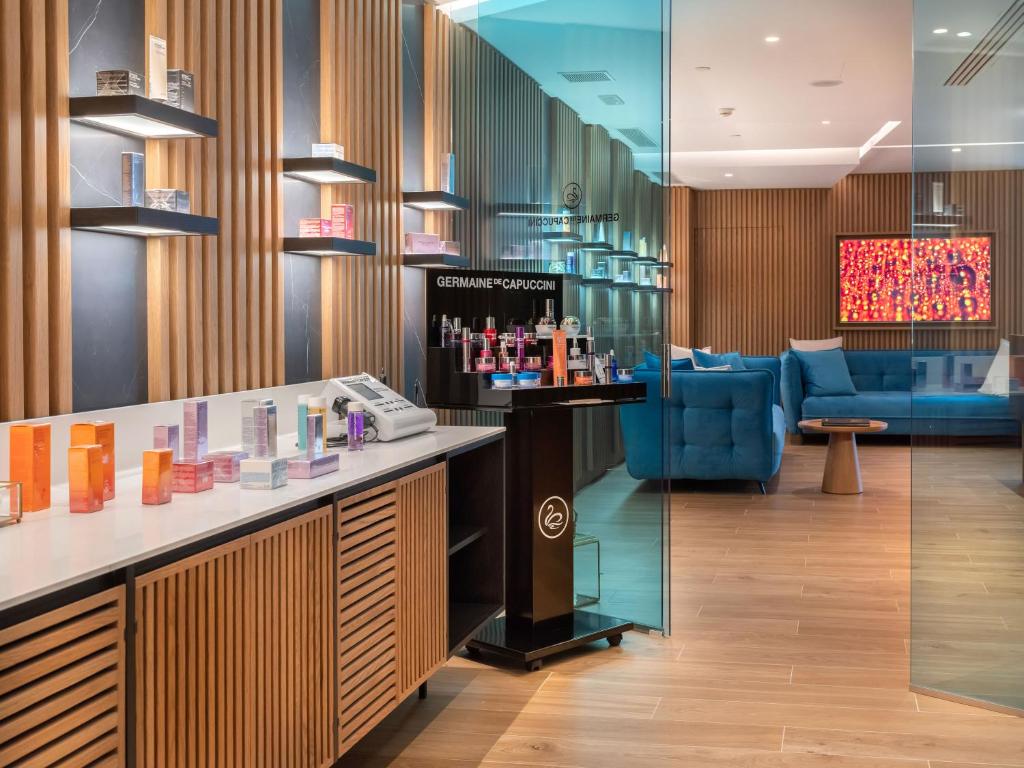 Mūsdienīgs skaistumkopšanas salons ar elegantu interjeru, kas ietver koka un akmens akcentus, un izstādītiem kosmētikas produktiem uz vitrīnām. Zila dīvāna zona nodrošina komfortu apmeklētājiem.