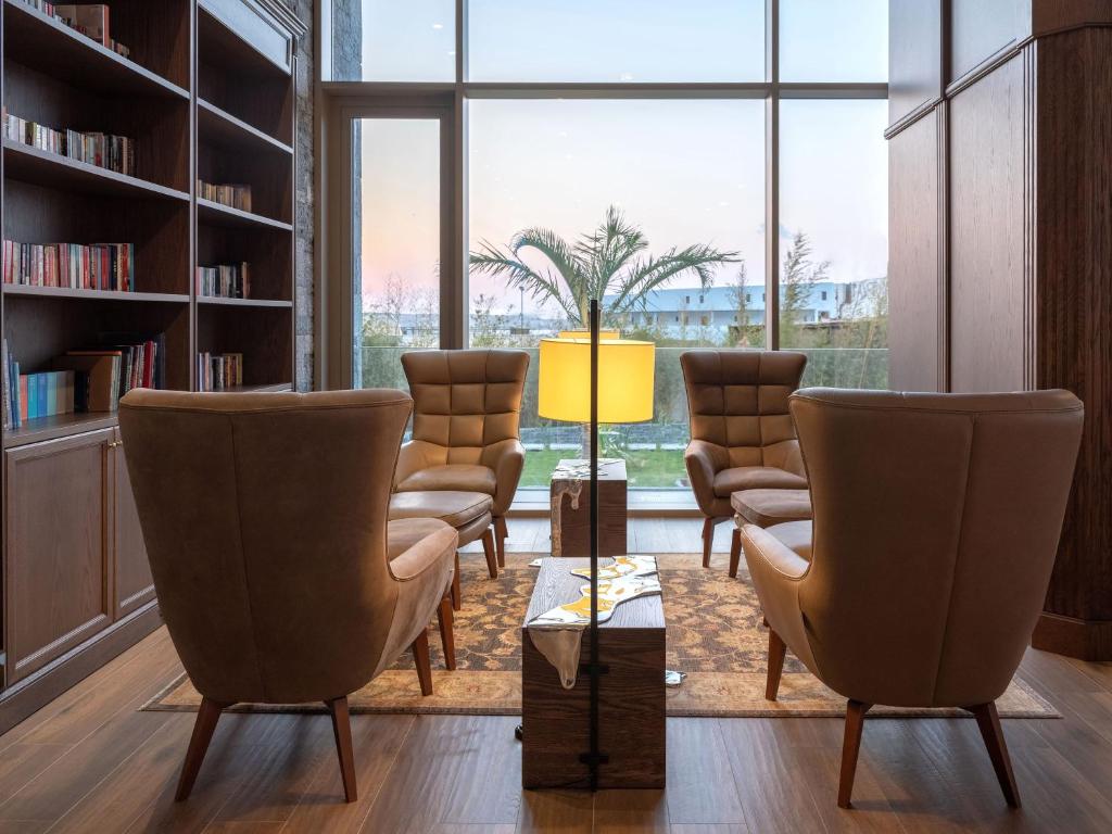 Mūsdienīga atpūtas telpa ar ērtām krēslām, grāmatu plauktu un logiem, caur kuriem redzams dārzs. Interjers ir apgaismots ar lampu un dekoratīvu paklāju.