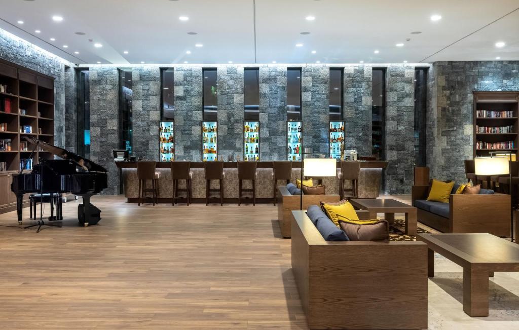 Mūsdienīgs viesnīcas vestibils ar klavierēm un bāru, kurā ir eleganti dīvāni un grāmatu plaukti. Telpa ir apgaismota ar mīkstu gaismu, radot patīkamu atmosfēru.