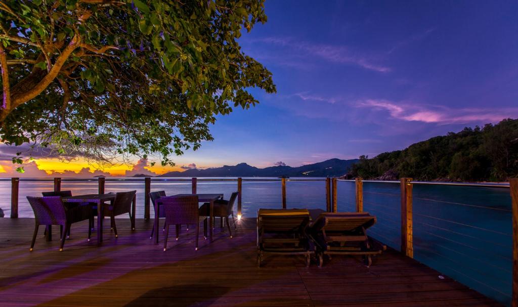 Romantiska saulrieta skatu uz okeānu ar terasi, kurā ir galdi un sauļošanās krēsli, apstādījumi un kalnu fons.