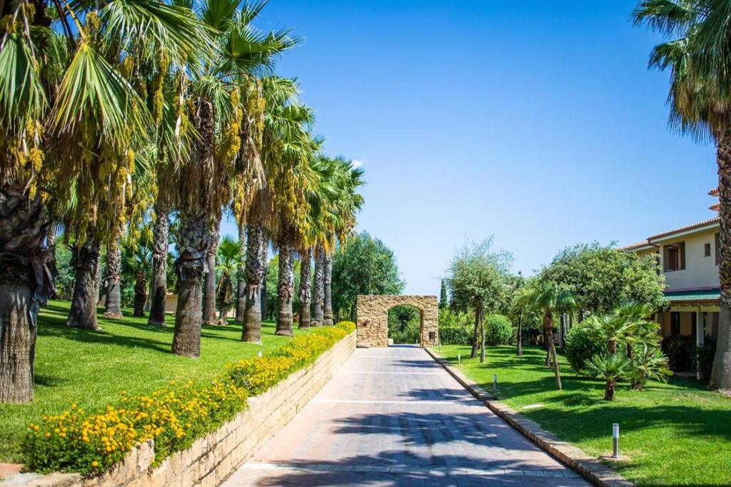 Skaista ceļa ainava, ko ieskauj palmas un ziedi, ved uz akmens vārtiem un ēkām. Zila debesis rada mierīgu atmosfēru.