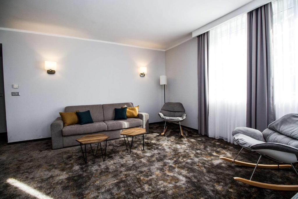 Mūsdienīga viesistaba ar pelēku dīvānu, diviem krēsliem un diviem koka galdiņiem, apgaismota ar siltu gaismu. Interjers ir vienkāršs un elegants, ar lieliem logiem un mīkstu paklāju.