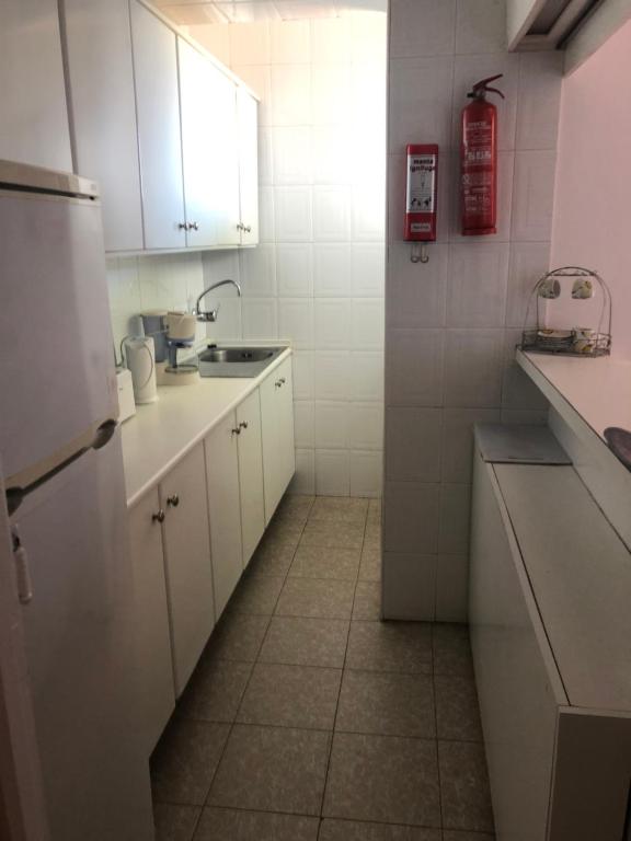 Kompaktā virtuve ar baltām skapēm un flīzēm, aprīkota ar izlietni un elektroierīcēm. Redzama ugunsdzēsības aparāts un gaismas avots.