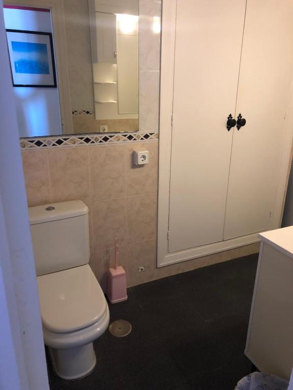 Tīra un gaiša vannas istaba ar tualeti, spoguli un baltu skapīti. Interjerā redzams rozā tualetes suku turētājs un flīžu apdare.