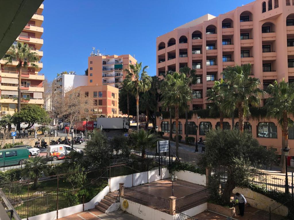 Skats uz pilsētas ainavu ar modernām ēkām, palmu kokiem un satiksmi. Gaismas apstākļi ir saulaini, radot dzīvīgu atmosfēru.