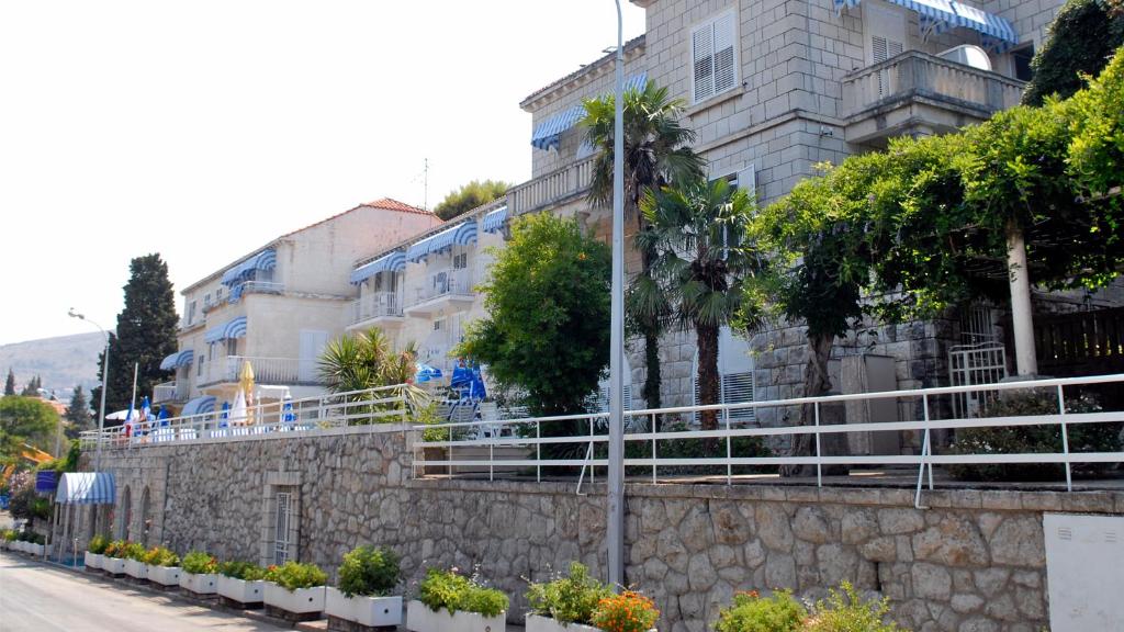 Vietējās arhitektūras ēka ar zilu un baltu stilu, apzaļumota ar palmām un krūmiem, atrodas blakus ceļam. Ap ēku ir izvietoti ziedu podi, kas piešķir skats patīkamu atmosfēru.