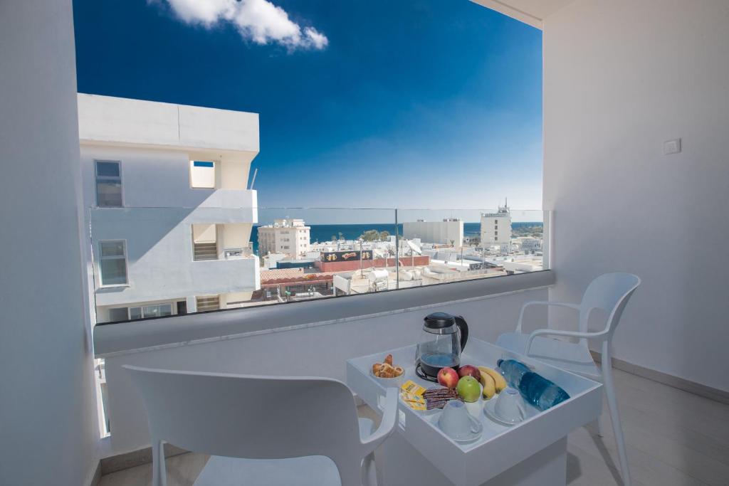 Balkons ar skatu uz pilsētu un jūru, uz galda atsvaidzinājumi un augļi. Mūsdienīga arhitektūra un gaiša atmosfēra.