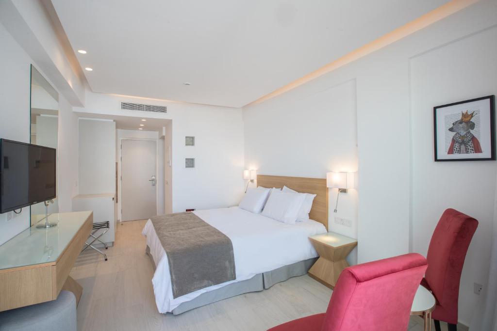 Mūsdienīga viesnīcas numura interjers ar ērtu divguļamo gultu, televizoru un sarkaniem krēsliem. Gaismas tonis un minimālistisks dizains rada relaksējošu atmosfēru.