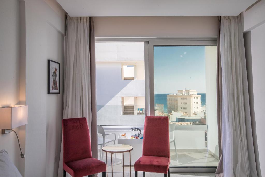 Mūsdienīgs viesnīcas numurs ar divām sarkanām krēsliem un skatu uz jūru. Logi ļauj ieplūst dabiskajai gaismai, radot patīkamu atmosfēru.