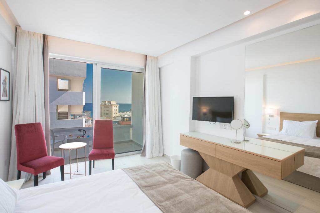 Mūsdienīgs viesnīcas numurs ar skatu uz jūru, ietver divus sarkanos krēslus un televizoru. Telpa ir gaiša un eleganta, ar lieliem logiem un minimālistisku interjeru.