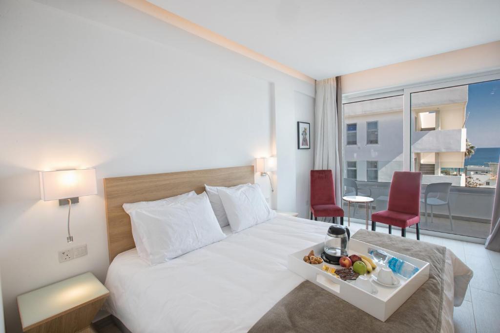 Mūsdienīgs viesnīcas numurs ar ērtu gultu, galdiņu un krēsliem, kā arī skatu uz jūru. Uz galda ir pasniegts brokastu paplāte ar augļiem un dzērieniem.