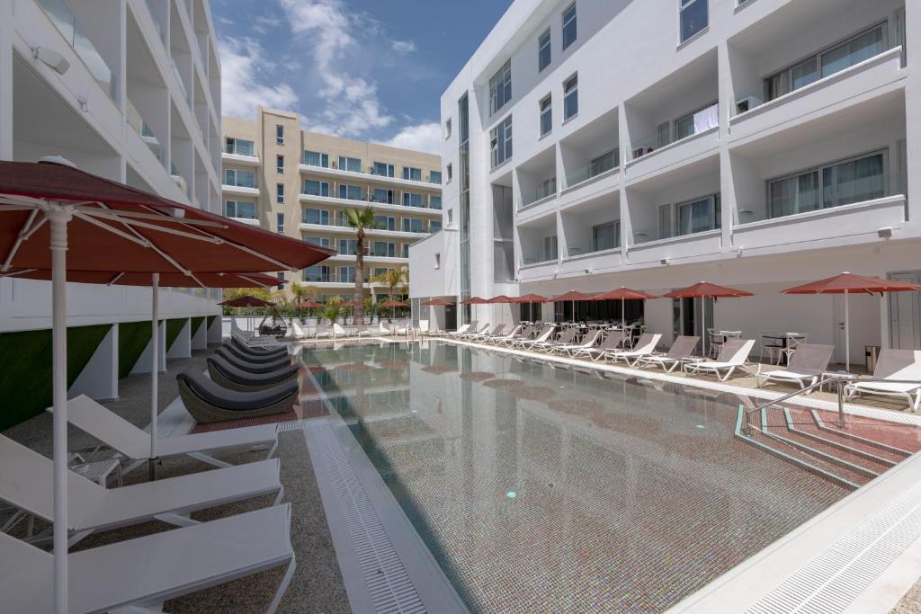 Mūsdienīgs viesnīcas baseins ar saulessargiem un sauļošanās krēsliem, kas atrodas starp baltiem ēku sienām un palmām. Ideāla vieta relaksācijai un atpūtai.