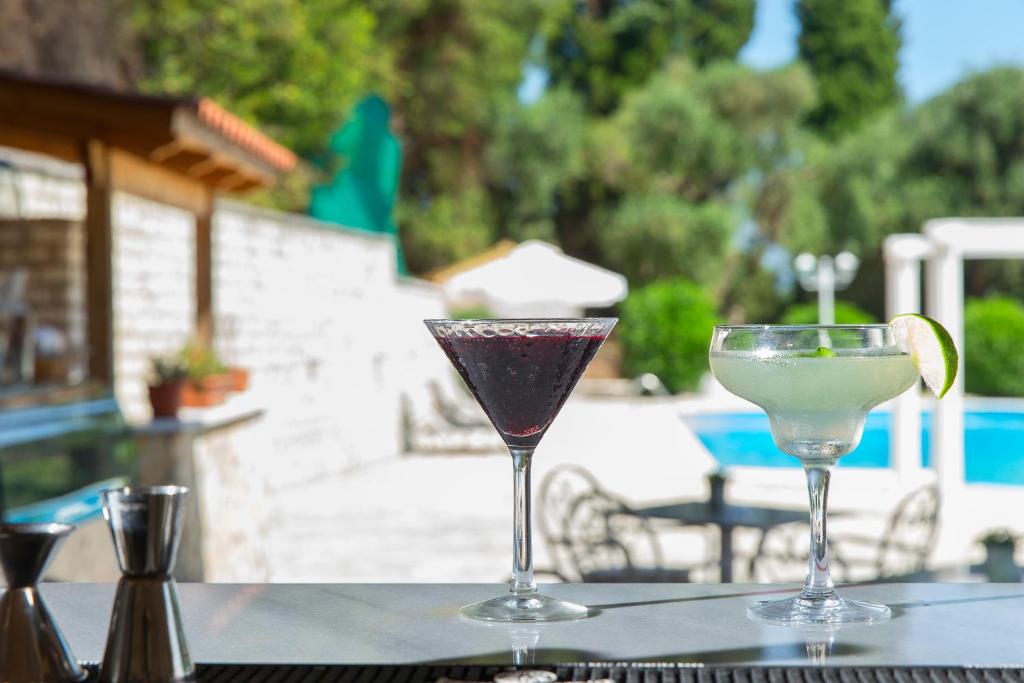 Divi kokteiļi, viens tumšs un viens zaļš, novietoti uz bāra letes ar peldbaseinu un dārzu fonā. Ideāla atmosfēra vasaras atpūtai.