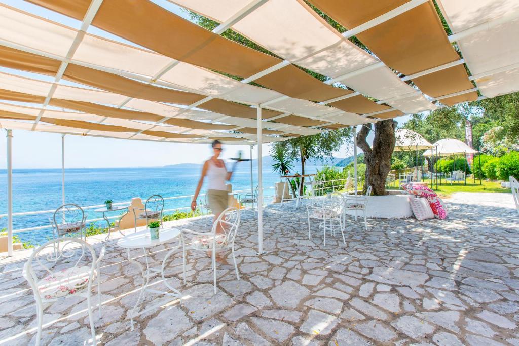 Tīra terase ar skatu uz jūru, kurā ir balti dzelzs krēsli un galdi, kā arī saulessargi. Fonā redzams zaļš zāles laukums un jūra, kas stiepjas tālumā.