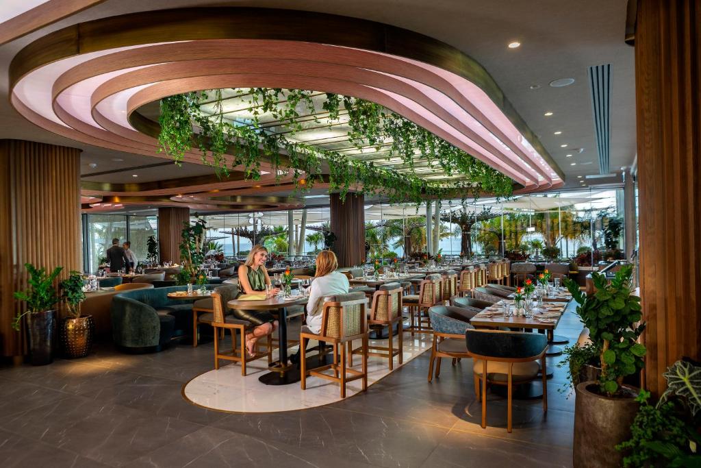 Mūsdienīgs restorāns ar elegantu interjeru, kas rotāts ar zaļumiem un modernām gaismām, radot relaksējošu atmosfēru. Apmeklētāji izbauda maltīti pie galda, ko ieskauj augi un stilīgas mēbeles.