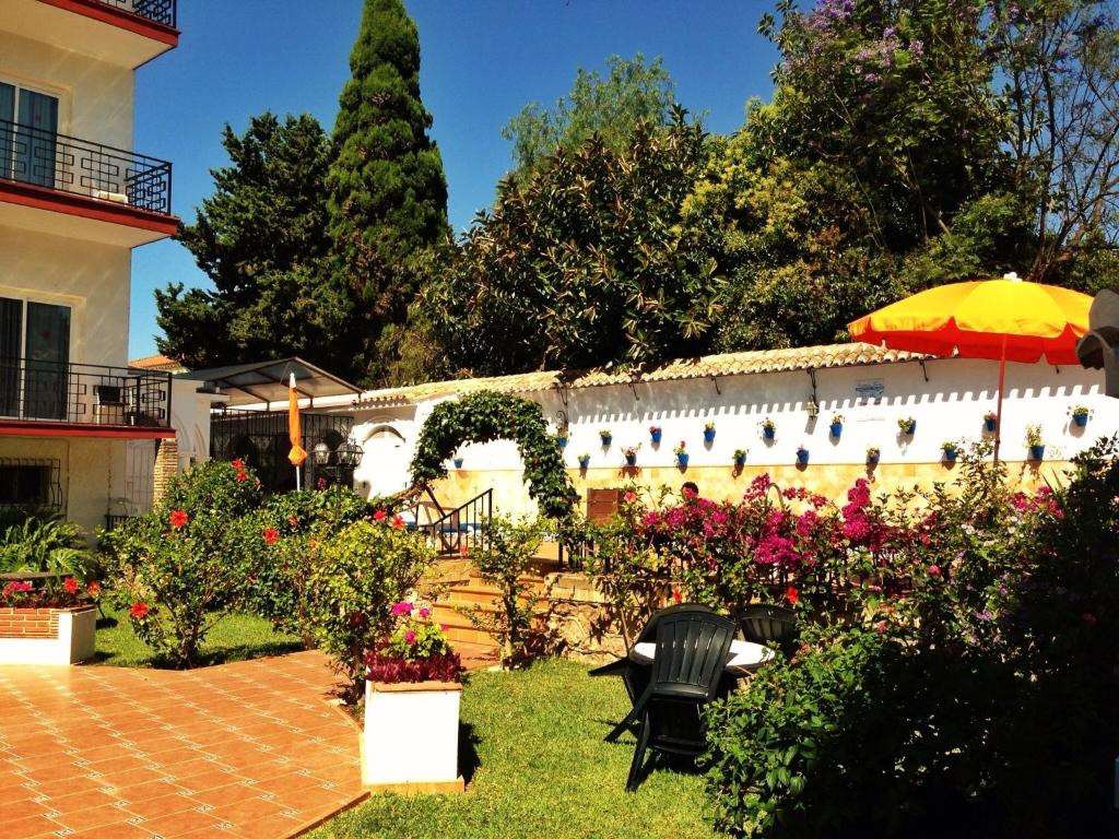 Dārzs ar krāsainiem ziediem un terasi, kurā ir sarkans galds un krēsli, ieskauts zaļām augiem un augstiem kokiem. Fons veido moderns, balts ēku komplekss ar balkoniem.