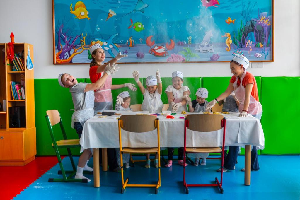 Bērni kopā ar pieaugušajiem jautri nodarbojas ar radošu aktivitāti, veidojot mākslu pie galda, kas ir klāts ar krāsainiem materiāliem. Fons ir krāsains jūras dzīves gleznojums, kas papildina atmosfēru.