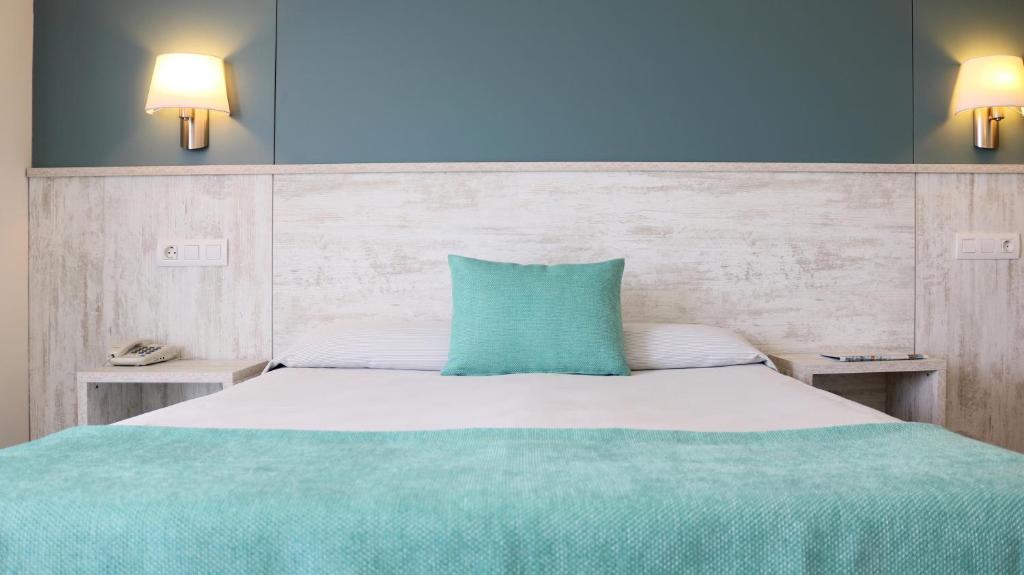 Mūsdienīgs guļamistabas interjers ar gaišām krāsām un stilīgu dizainu, izceļot gultu ar zilu sega un spilvenu. Sienas apgaismojums un koka akcenti piešķir telpai siltumu un komfortu.