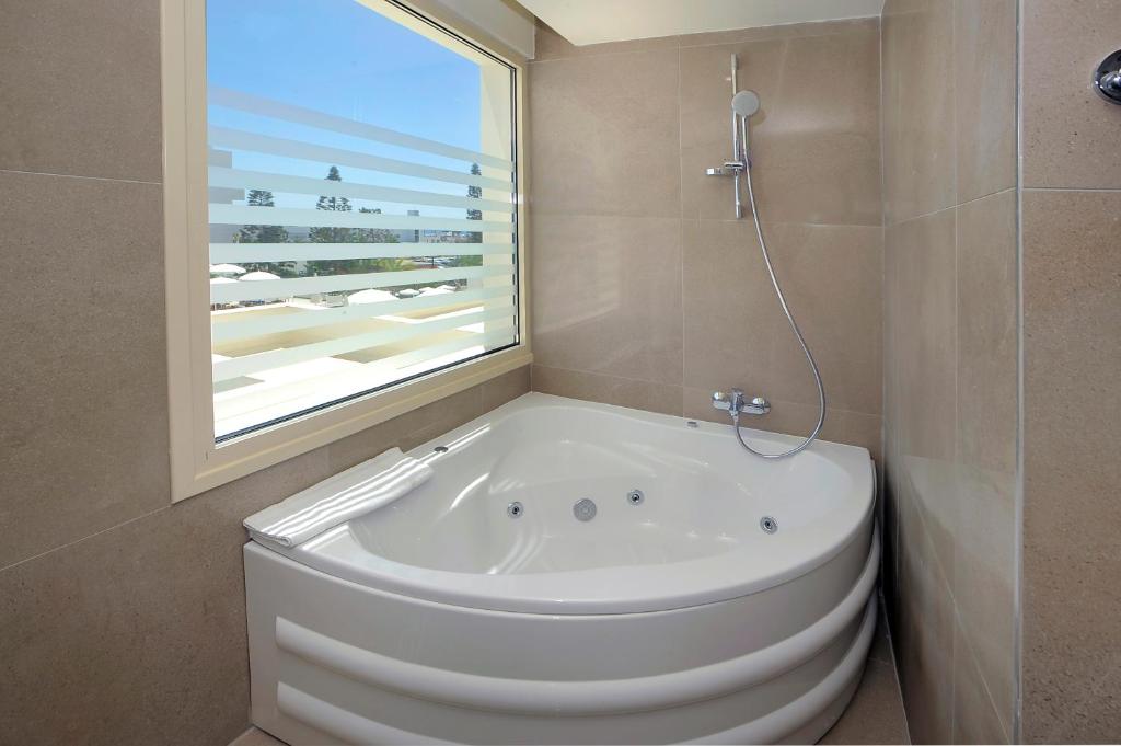 Mūsdienīgs vannas istabas stūris ar balto džakuzi un lieliem logiem, kas piedāvā skatu uz ārpusi. Sienas ir klātas ar gaišām flīzēm, radot elegantu un relaksējošu atmosfēru.