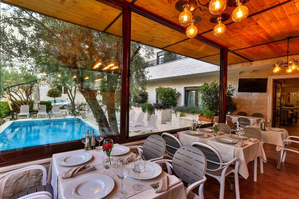 Eleganti iekārtota restorāna iekštelpa ar galdiem un skatu uz baseinu, kas ieskauts zaļumā. Gaismas instalācija pie griestiem piešķir siltu atmosfēru.