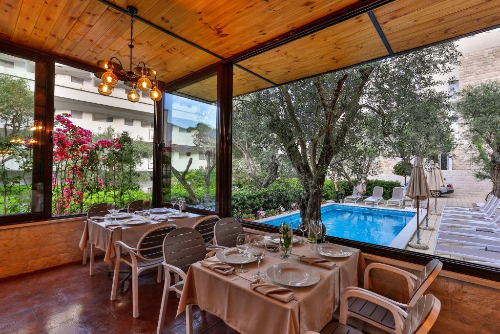 Restorāna interjers ar galdiem, kas izvietoti blakus logiem, caur kuriem ir redzams dārzs un baseins. Apkārtne ir zaļa un ziedējoša, radot relaksējošu atmosfēru.