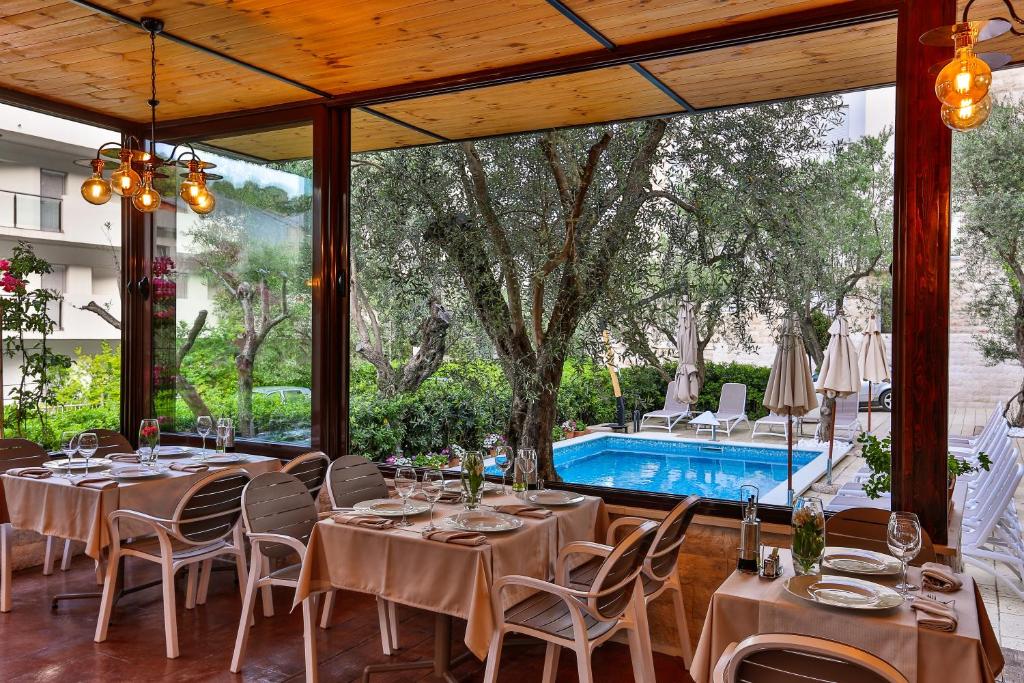 Gaiša un eleganta restorāna iekārtojums ar galdiem un krēsliem, skats uz peldbaseinu un dārzu caur lieliem logiem. Ideāla vieta relaksācijai un maltītēm brīvā gaisotnē.