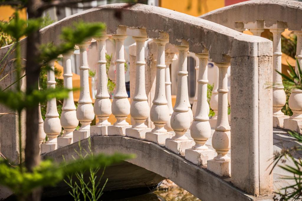 Balts tilts ar dekoratīvām margām, kas ieskauj dārza ainavu. Tilts rada mierīgu atmosfēru un piešķir šarmu apkārtējai videi.