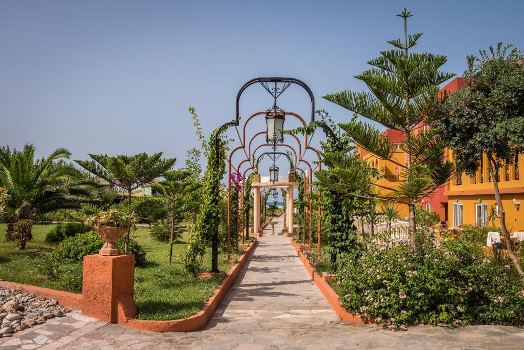 Skaists dārza ceļš ar dekoratīvām arkas un zaļumiem, ko ieskauj sulīgas palmas un ziedi. Fons veido krāsaini ēku fasādes.