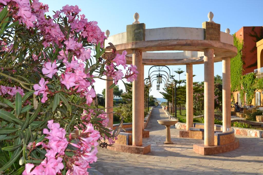 Krāšņs dārzs ar rozā ziediem un arhitektūras elementiem, kas ietver kolonnā veidotu paviljonu un ainavisku ceļu. Ideāla vieta relaksācijai un dabas baudīšanai.