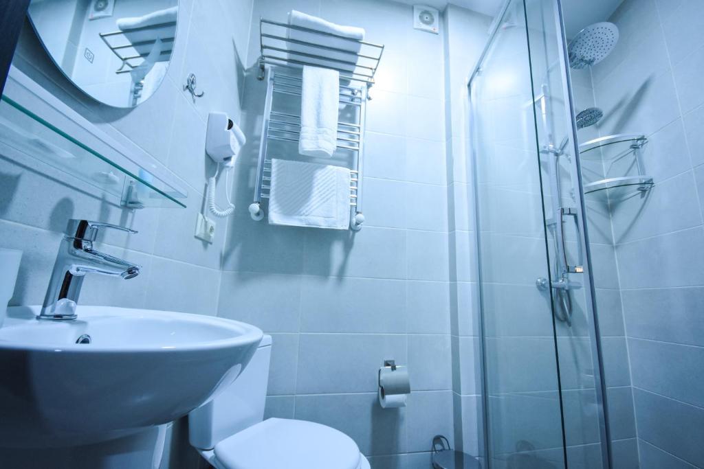 Mūsdienīga vannas istaba ar stikla dušu, balto izlietni un higiēnas piederumiem. Telpa ir gaiša ar pelēkām flīzēm un elegantu dizainu.