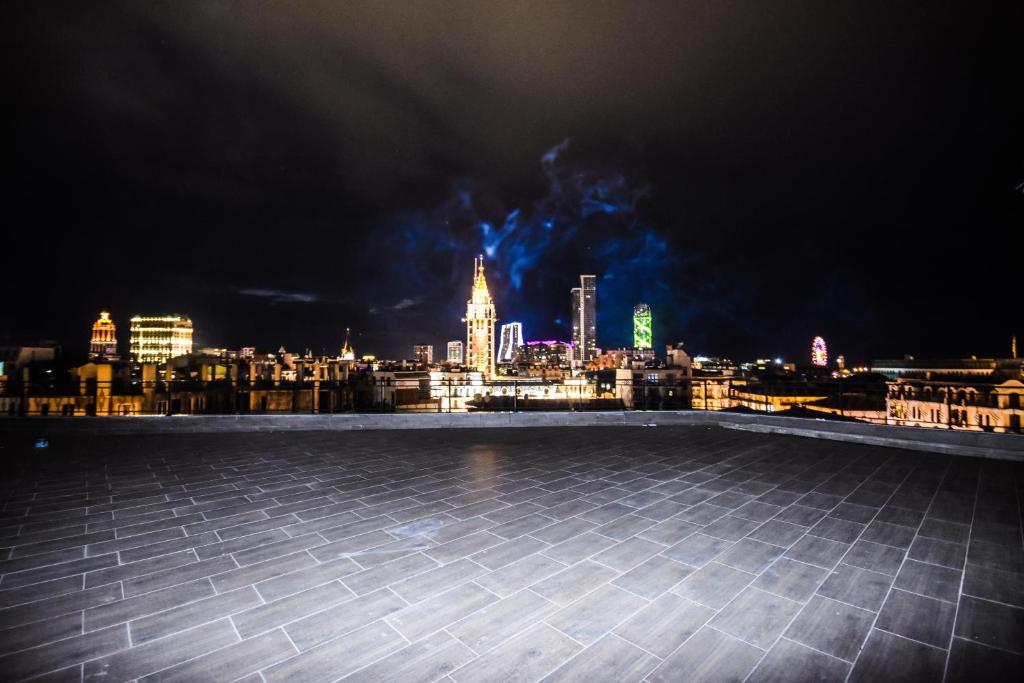 Nakts skats uz pilsētu ar modernām ēkām un krāsainām gaismām. Jumta terase ar flīzēm un tumšu debesu fona.