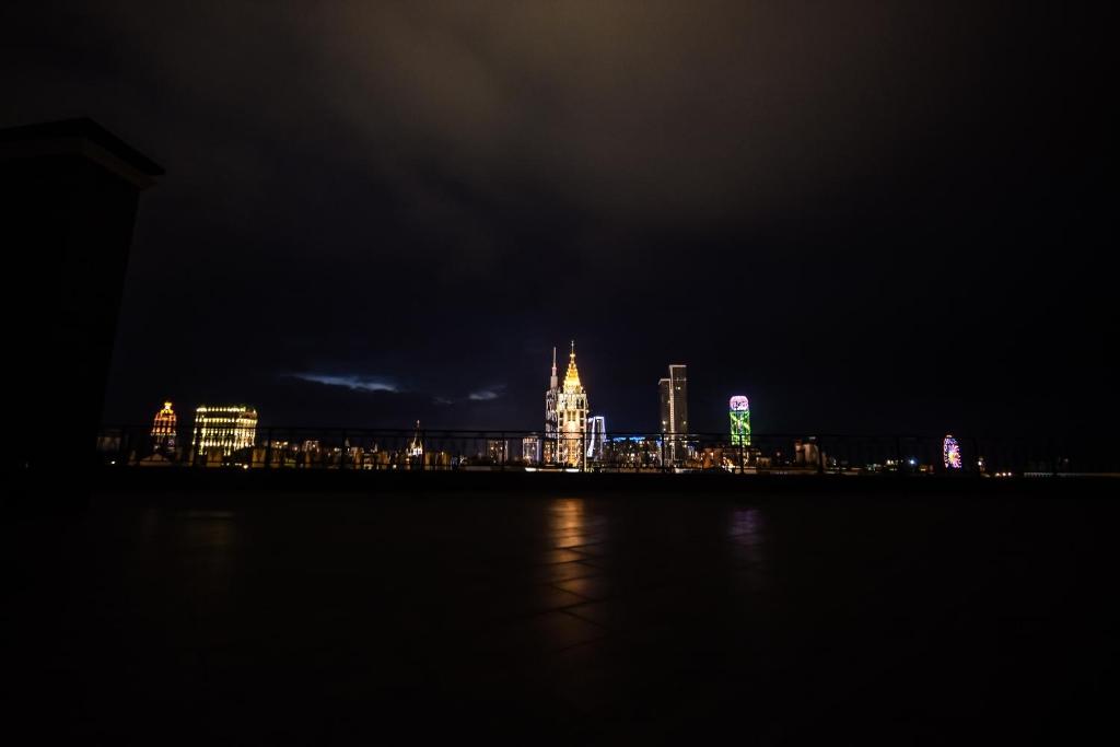 Nakts ainava ar apgaismotiem debesskrāpjiem un spīdīgu pilsētas panorāmu, kas rada noslēpumainu atmosfēru.