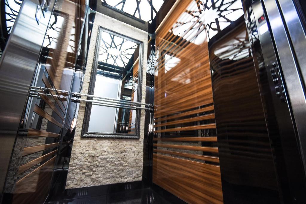 Mūsdienīgs lifts ar spoguļotām un koka detaļām, kas rada elegantu un stilīgu atmosfēru. Interjerā ir redzams liels spogulis un dekoratīva griestu apgaismojuma konstrukcija.