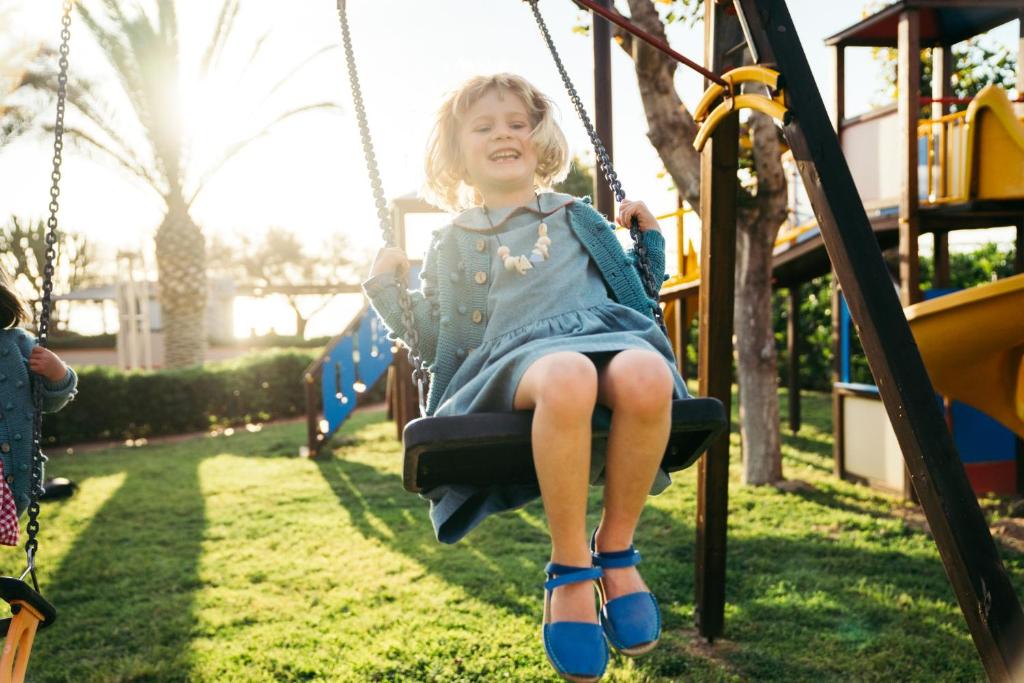 Bērns priecīgi šūpojas šūpolēs parkā, fonā redzami palmām un rotaļu laukums. Saules gaisma izgaismo ainu, radot laimīgu atmosfēru.