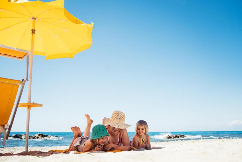 Pludmales aina ar saulainiem bērniem un pieaugušajiem, kas atpūšas zem dzeltena saulessarga. Pavasarīga atmosfēra ar zilu debesu fonu un mierīgu jūru.