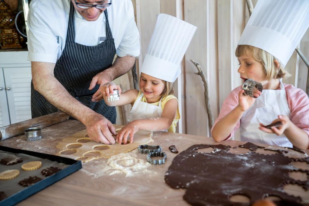 Bērni un pieaugušais kopā cep kūkas virtuvē, izmantojot cepamo mīklu un formas. Uz galda redzama miltu un mīklas sajaukums, kā arī cepšanas piederumi.