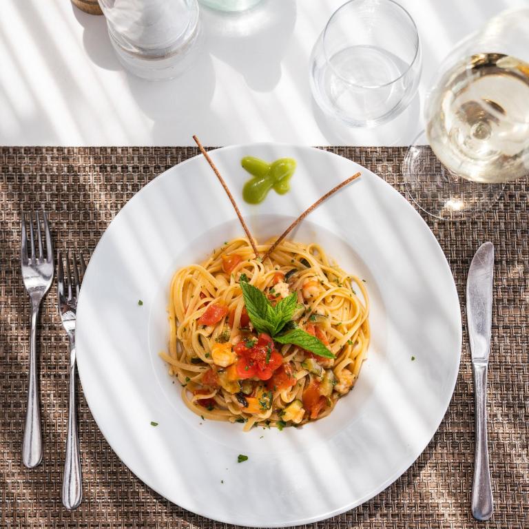 Pasta ar tomātiem un garšaugiem uz baltā šķīvja, dekorēta ar piparmētru lapām un kraukšķīgām nūjiņām, pasniegta uz galda ar ūdens glāzēm un vīna glāzi.