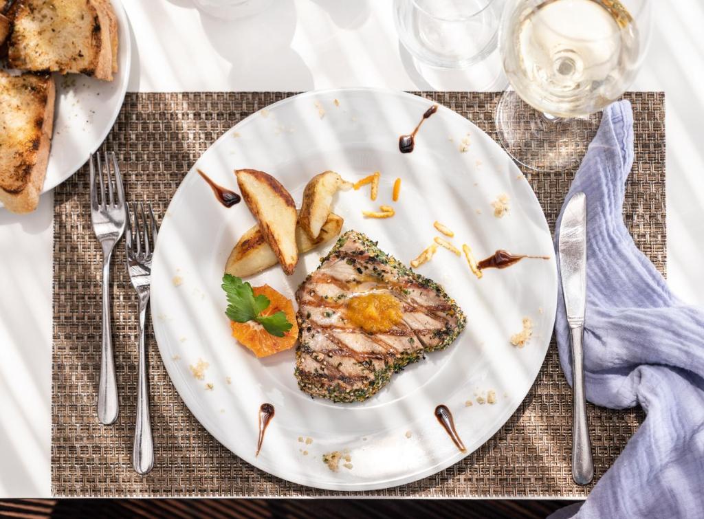 Garšīgs cepts zivs filejas gabals ar kartupeļu šķēlītēm un apelsīnu, pasniegts uz baltas šķīvja, dekorēts ar zālītes un mērci. Blakus atrodas grauzdiņi un glāze vīna.