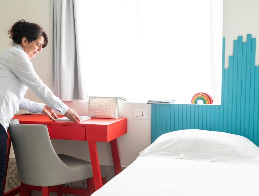 Sieviete sakārto galdu modernā istabā ar sarkanu galdu un gaišām krāsām. Logā ieplūst dabiskais apgaismojums, radot patīkamu atmosfēru.