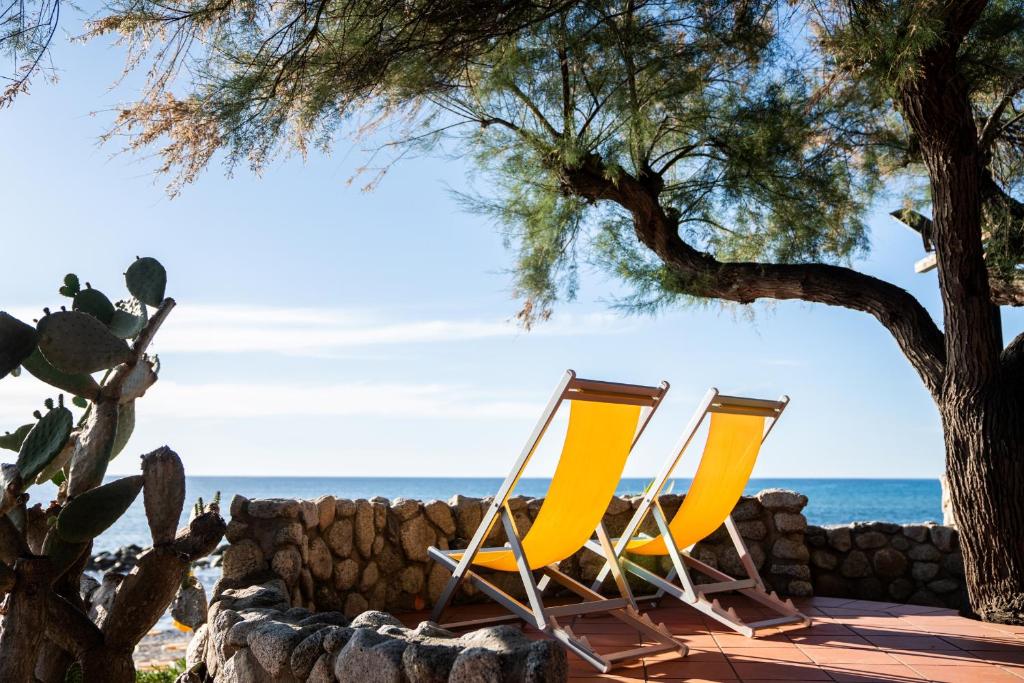 Saulainas pludmales krēslus ar dzeltenām sēdvietām, kas izvietoti pie akmens sienas, ar skatu uz jūru un koku ēnu. Ideāla vieta atpūtai pie jūras.