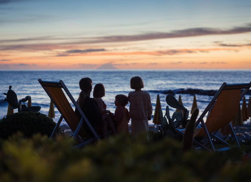 Bērni skatās saulrietu pie jūras, sēžot uz krēsliem. Ūdens un debesu krāsas rada mierīgu un burvīgu atmosfēru.