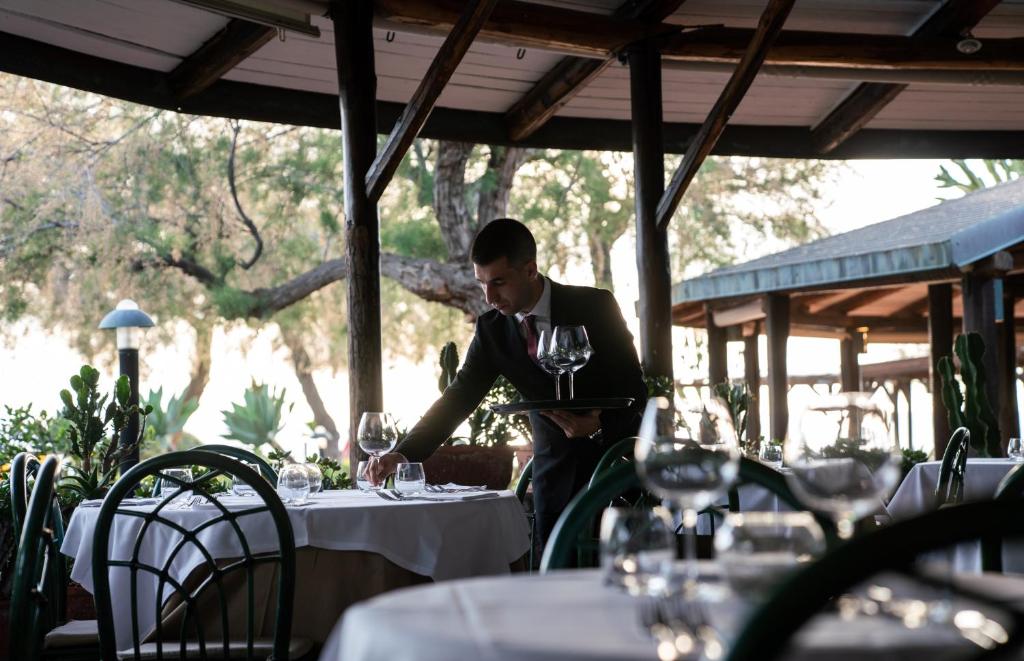 Galdnieks, tērpies uzvalkā, pasniedz vīna glāzes elegantā restorānā ar koka konstrukcijām un zaļiem augiem. Gaisotne ir mierīga un gaida viesus.