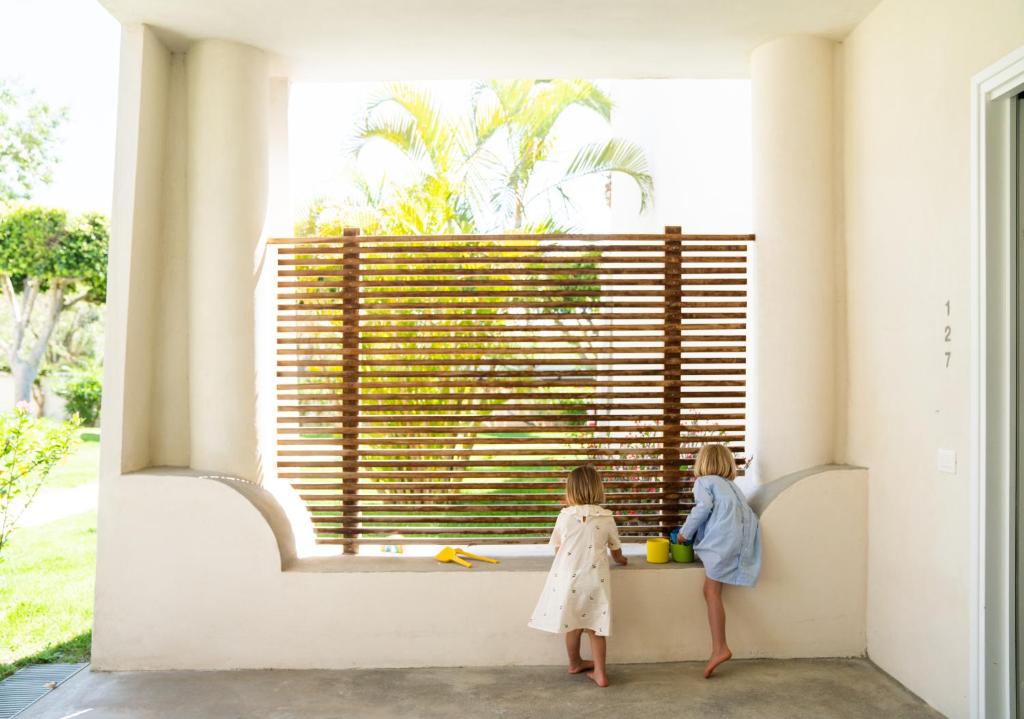 Divas meitenes rotaļājas ar krāsainām traukiem saulainā terasē, kamēr aiz viņām redzams zaļš dārzs. Gaismas plūsma caur koka žogu rada mājīgu atmosfēru.