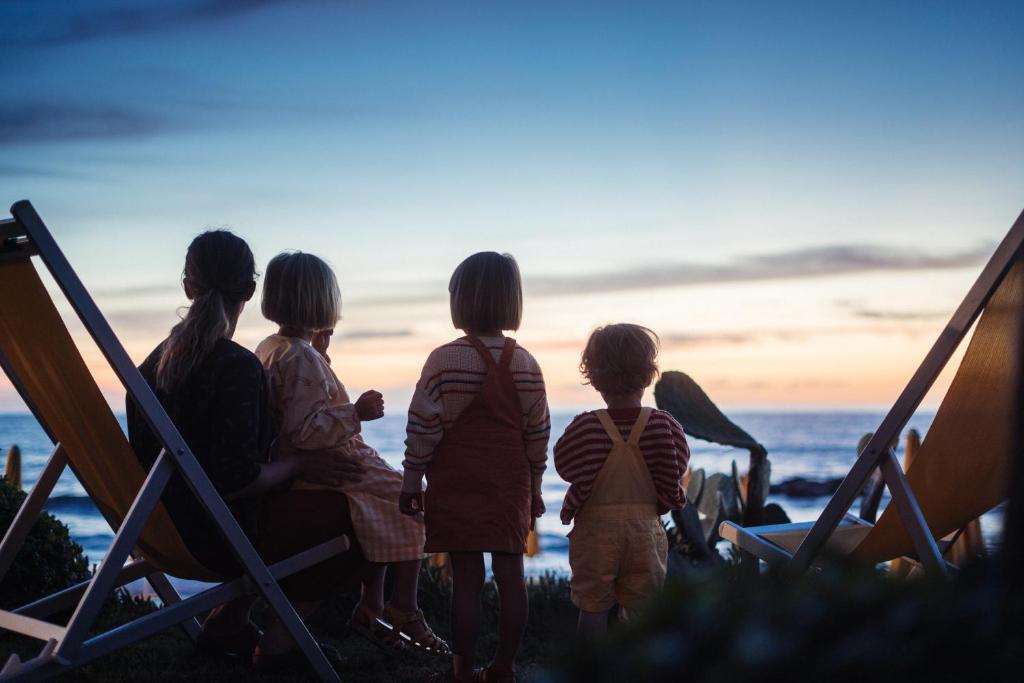 Bērni skatās saulrietu pie jūras, sēžot uz šūpuļkrēsliem. Mierīga un nostalģiska atmosfēra.