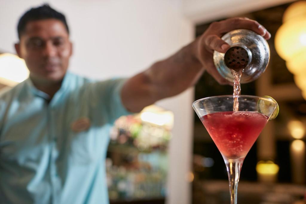 Bārmenis ielej sarkano kokteili martini glāzē, pievienojot citrona šķēlīti. Fona apgaismojums rada siltu un viesmīlīgu atmosfēru.