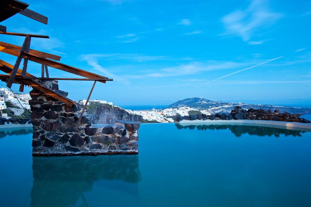 Skaists skats uz baseinu ar koka konstrukciju, kas sniedzas virs ūdens, un panorāmas skatu uz Santorini salas ainavu.