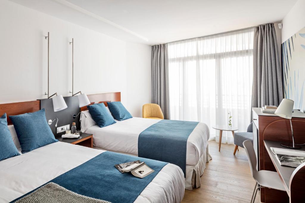 Mūsdienīgs viesnīcas numurs ar divām gultām, dekorēts ar zilu un baltu krāsu, ērtām mēbelēm un lieliem logiem, kas nodrošina dabisko apgaismojumu.