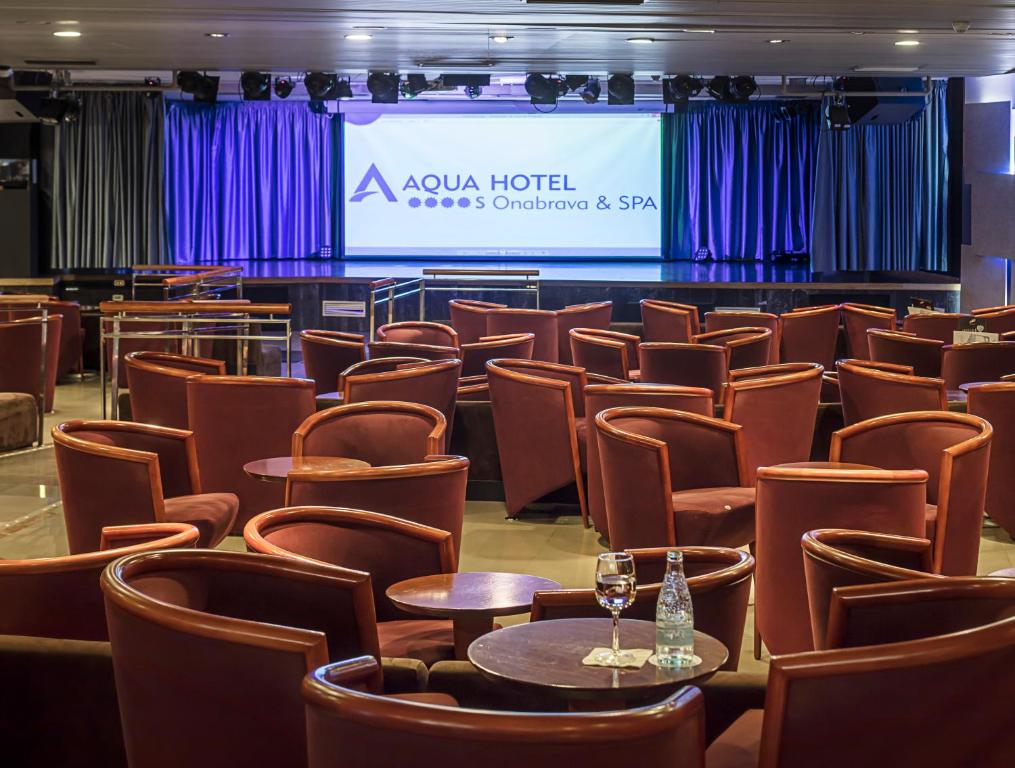 Aqua Hotel Onabrava & SPA pasākumu zāle ar elegantiem krēsliem un galdiņiem, gatava viesu uzņemšanai. Skatuves fons ar zilu aizkaru un projektoru ekrānu piedāvā ideālu vidi pasākumiem.